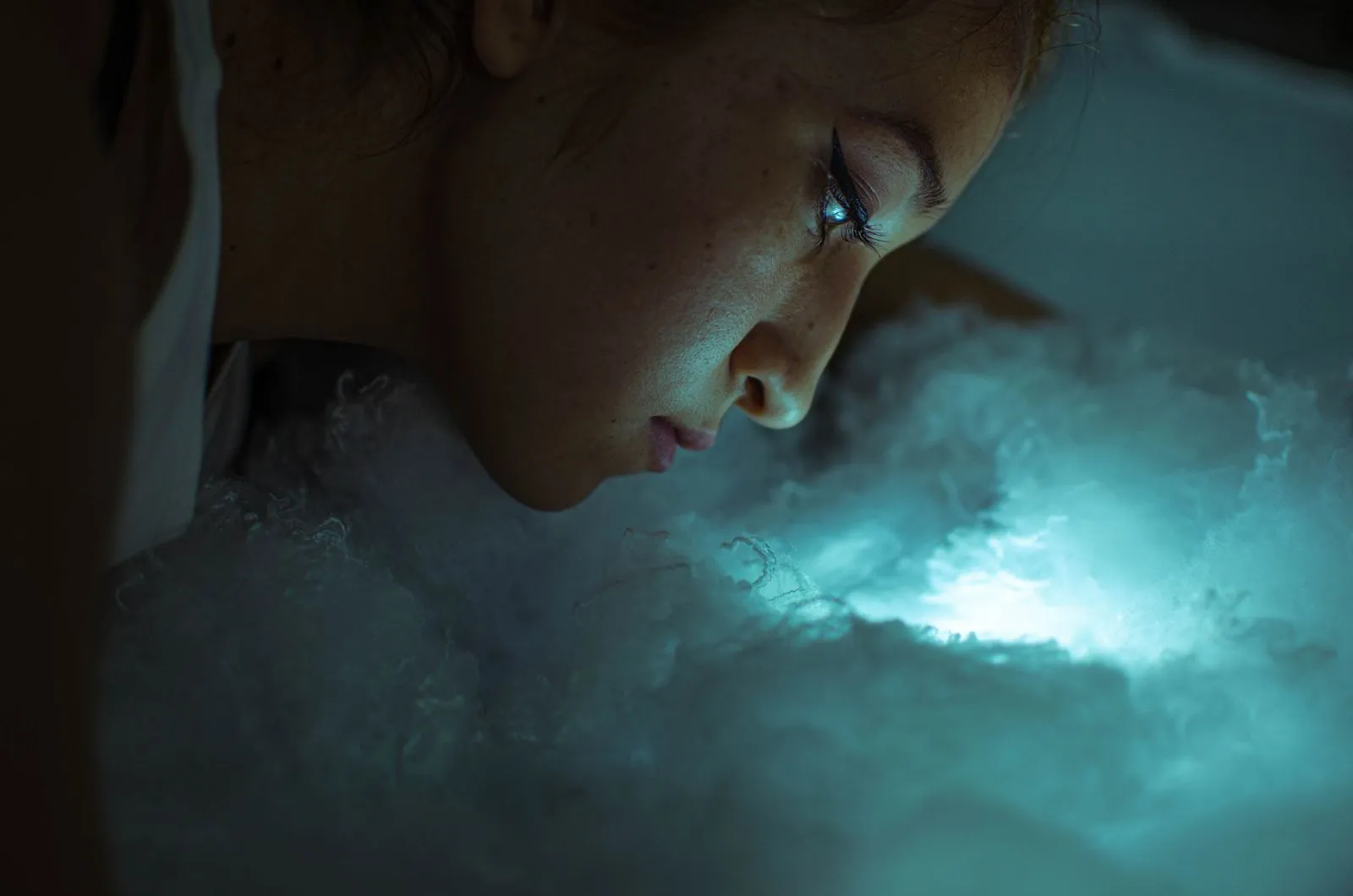 Close-up of a woman’s side profile illuminated by ethereal blue-green light resembling clouds.
